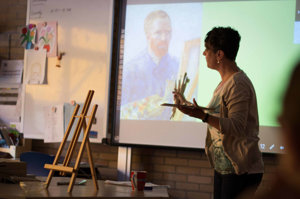 Een onderwijzeres met penselen in haar hand staat voor het digibord waarop het schilderij Zelfportret als schilder is te zien. Foto: Brenda Roos