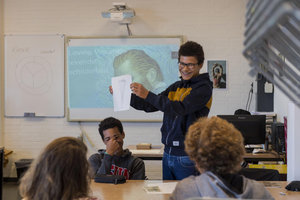 A boy with dark curls and glasses laughs and shows his assignment to the class. In the background the IWB with an image from the movie Loving Vincent. Photo: Jan-Kees Steenman