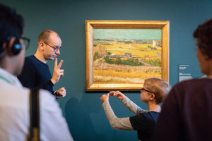 Een kind en een rondleider communiceren met elkaar in gebarentaal bij Van Goghs schilderij De oogst. Foto Brenda Roos